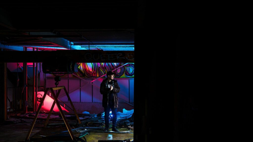 A person stands in a dimly lit, abandoned space with scattered debris. The scene is illuminated by vibrant neon lights in hues of blue and pink, casting a colorful glow on the graffiti-covered walls. The right side of the image is shrouded in darkness. Image by Documentary Wedding Photographer Lewis Bishop Photography.