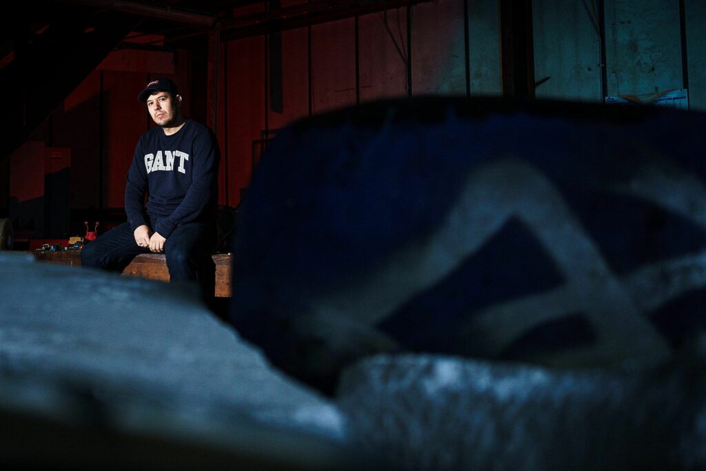 A man wearing a dark baseball cap and a sweatshirt with the word "SAINT" sits on a wooden bench in a dimly lit industrial space. The foreground is blurred with an indistinguishable object, and the background features metal beams with red and blue lighting. Image by Documentary Wedding Photographer Lewis Bishop Photography.