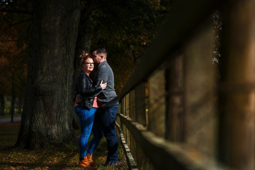 A couple stands closely together near a wooden fence in a park. The woman, with red hair in a bun and wearing glasses, a black jacket, and jeans, smiles and looks at the camera. The man, wearing a grey jacket and jeans, leans in close, facing her. Trees surround them. Image by Documentary Wedding Photographer Lewis Bishop Photography.