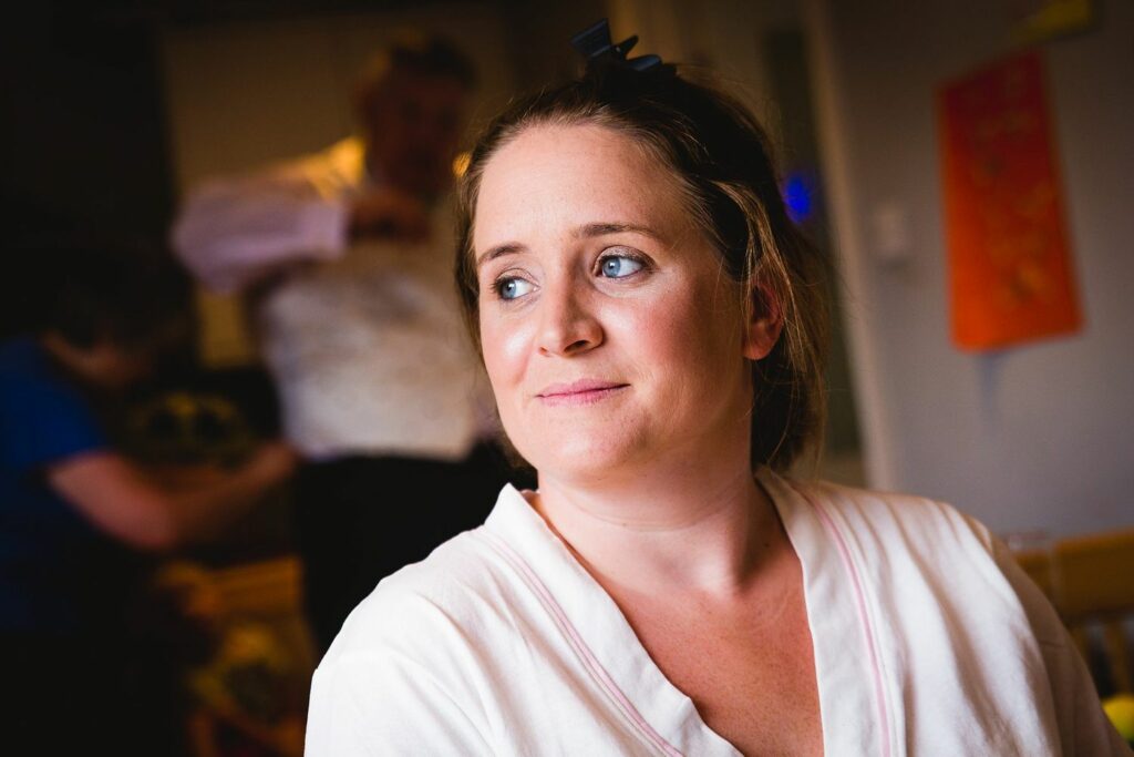 A woman with light brown hair and a black bow hair accessory gazes to her right with a soft smile. She wears a white top and is indoors. The background shows blurred figures of a man and another person involved in some activity. Image by Documentary Wedding Photographer Lewis Bishop Photography.
