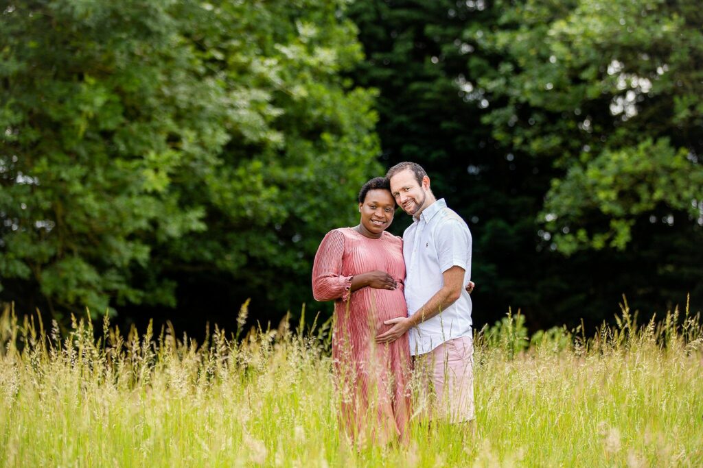 A pregnant woman in a pink dress and a man in a white shirt stand in a grassy field, smiling and embracing each other. The man is gently touching the woman's belly. The background features green trees and a vibrant, natural setting. Image by Documentary Wedding Photographer Lewis Bishop Photography.