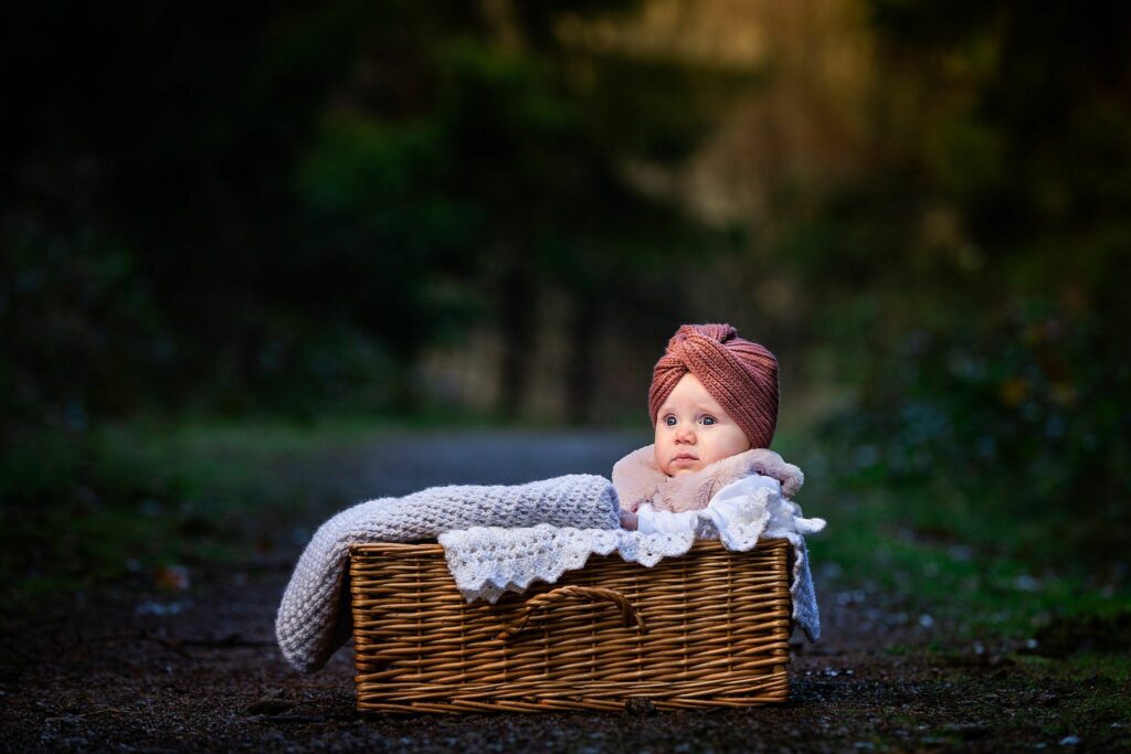 A baby wearing a pink turban headwrap and cozy clothing is lying in a large wicker basket outdoors. The basket is lined with a soft, white blanket. The background features a blurred natural setting with trees and greenery. The baby appears peaceful and content. Image by Documentary Wedding Photographer Lewis Bishop Photography.