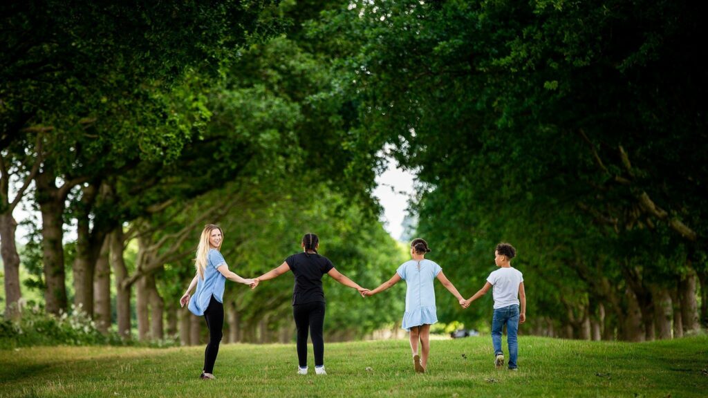 Four people, including two adults and two children, walk hand in hand down a tree-lined path. The adults are on the ends, with the children in the middle. The lush, green trees form a beautiful natural tunnel overhead, and everyone is walking away from the camera. Image by Documentary Wedding Photographer Lewis Bishop Photography.