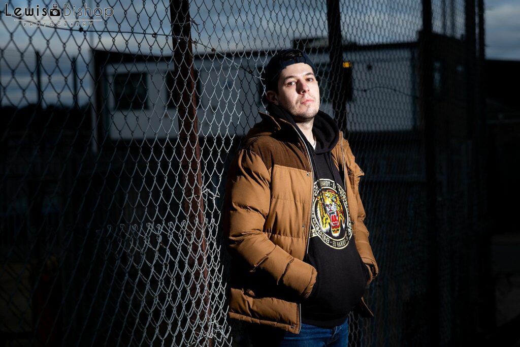 A person wearing a brown jacket and black hoodie with a tiger emblem leans against a chain-link fence. The individual has a serious expression and is outdoors, with buildings visible in the background. The sky is partly cloudy. Image by Documentary Wedding Photographer Lewis Bishop Photography.
