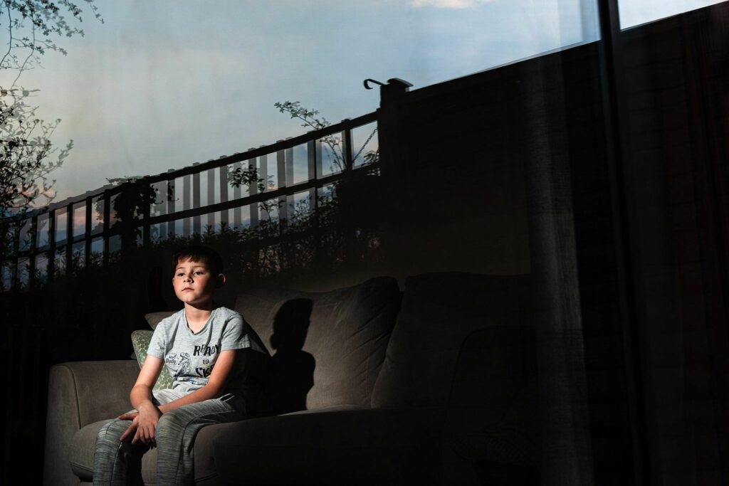 A boy sits on a couch in a dimly lit room, gazing thoughtfully out a window. The reflection of an outdoor scene with trees and a fence is visible in the glass, blending with the indoor view and casting soft shadows on the boy and the interior. Image by Documentary Wedding Photographer Lewis Bishop Photography.