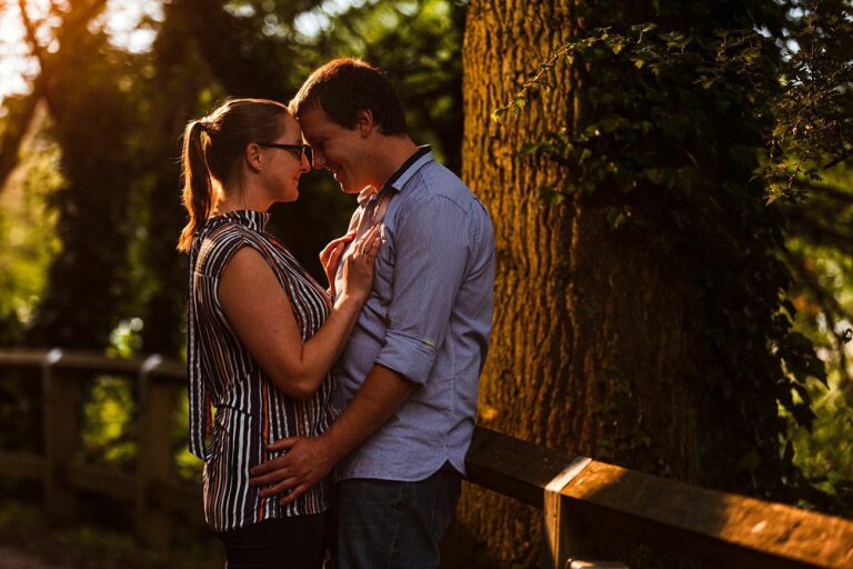 A couple stands close together on a sunlit path, embracing and sharing a tender moment. They are surrounded by greenery, with large trees and vines in the background. The warm light creates a romantic and intimate atmosphere. Both are smiling and wearing casual clothes. Image by Documentary Wedding Photographer Lewis Bishop Photography.