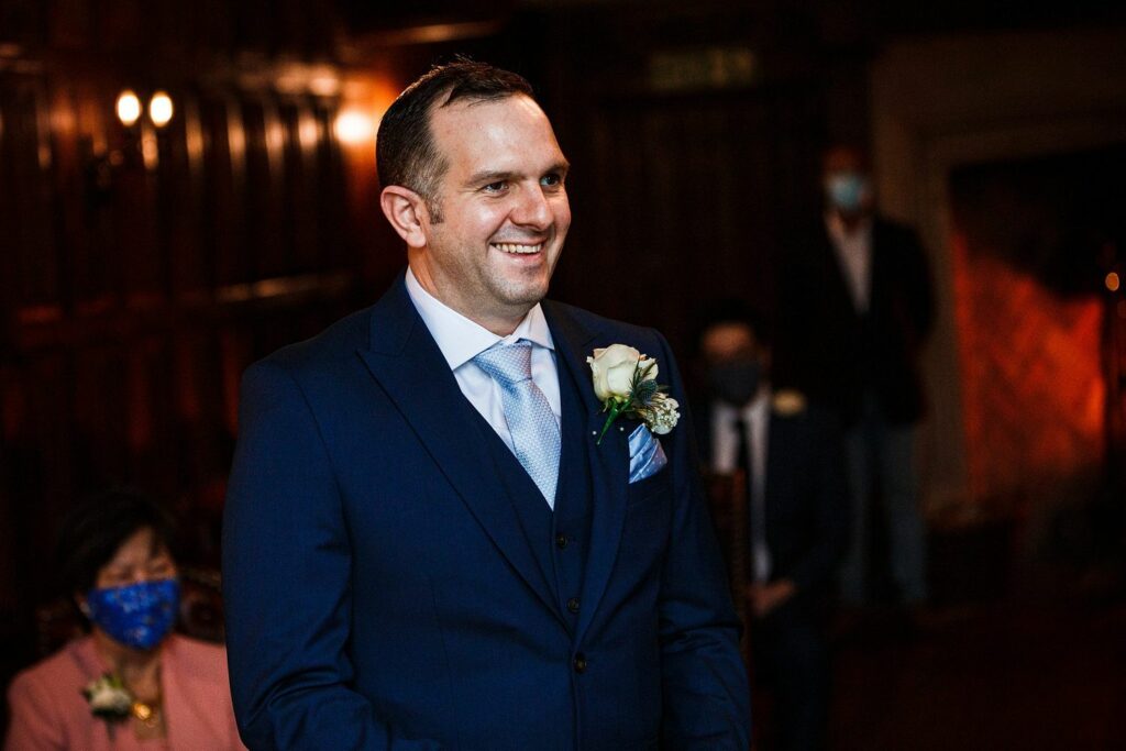 A man in a navy blue suit, light blue tie, and white shirt, smiles while standing indoors. He has a boutonniere with white flowers on his left lapel. The background shows wooden paneling, people wearing masks, and a lit fireplace. Image by Documentary Wedding Photographer Lewis Bishop Photography.