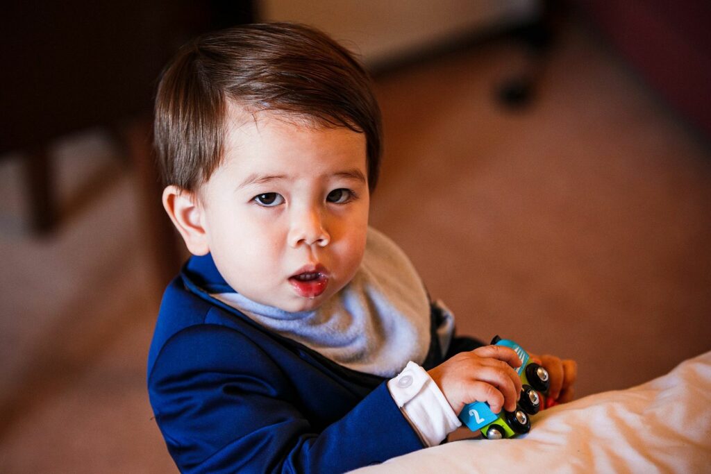 A young child with light skin and dark brown hair, dressed in a blue suit and white shirt, holds a small blue toy train. The child looks towards the camera with a curious expression. The background is softly blurred, focusing attention on the child. Image by Documentary Wedding Photographer Lewis Bishop Photography.