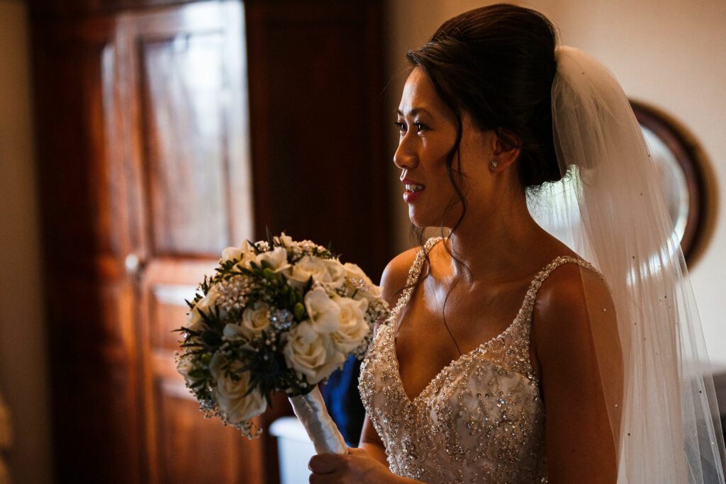 A bride stands in a warmly lit room, holding a bouquet of white flowers. She is wearing an intricately beaded wedding dress and a white veil. Her hair is styled in an elegant updo, and she has a content expression on her face. A wooden wardrobe and a circular mirror are in the background. Image by Documentary Wedding Photographer Lewis Bishop Photography.