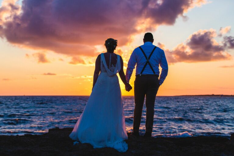 A bride and groom stand hand in hand, facing the ocean during sunset. The bride wears a flowing white dress, and the groom wears a white shirt with suspenders. The sky displays vibrant hues of orange, pink, and purple, with clouds scattered above the horizon. Image by Documentary Wedding Photographer Lewis Bishop Photography.