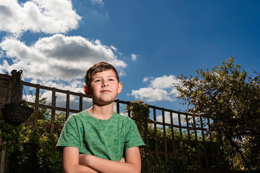 A young boy stands outside, arms crossed and looking off into the distance. He wears a light green shirt, and behind him is a wooden fence and lush greenery. The sky is bright blue with scattered white clouds, suggesting a sunny day. Image by Documentary Wedding Photographer Lewis Bishop Photography.