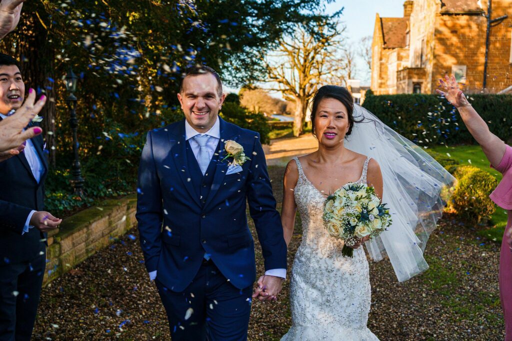 A newlywed couple walks hand in hand outdoors on a sunny day. The bride wears a white lace gown and veil, holding a bouquet of white roses. The groom is in a navy blue suit with a white boutonniere. Confetti is being thrown by smiling guests surrounding them. Image by Documentary Wedding Photographer Lewis Bishop Photography.