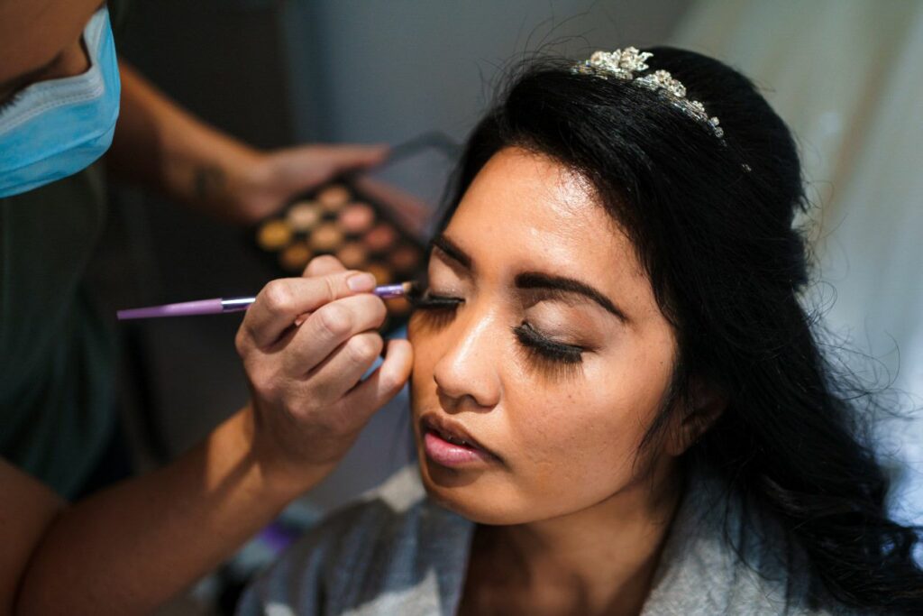 A woman with dark hair is sitting with her eyes closed while a makeup artist applies eye shadow to her eyelids using a brush. She is wearing a tiara and appears to be preparing for a special occasion. The makeup artist is wearing a mask. Image by Documentary Wedding Photographer Lewis Bishop Photography.