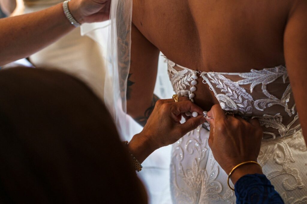 A bride in a white lace wedding dress is shown from the back as someone helps button her gown. The intricate floral lace design and the veil are visible, and several hands are seen assisting with the buttons. The scene captures a moment of wedding preparation. Image by Documentary Wedding Photographer Lewis Bishop Photography.