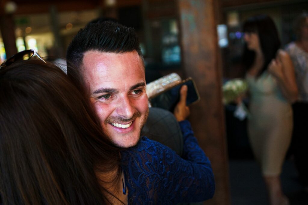 A man with short dark hair and a trimmed beard smiles broadly as he hugs someone. He is wearing a blue outfit. In the background, two women are seen; one holding a bouquet and another holding what appears to be a clutch. The setting seems to be indoors. Image by Documentary Wedding Photographer Lewis Bishop Photography.