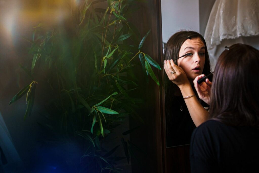 A woman is standing in front of a mirror, applying mascara to her eyelashes. She is holding the mascara wand close to her eye while looking into the mirror. There is a leafy green plant and a white piece of clothing visible in the background. Image by Documentary Wedding Photographer Lewis Bishop Photography.