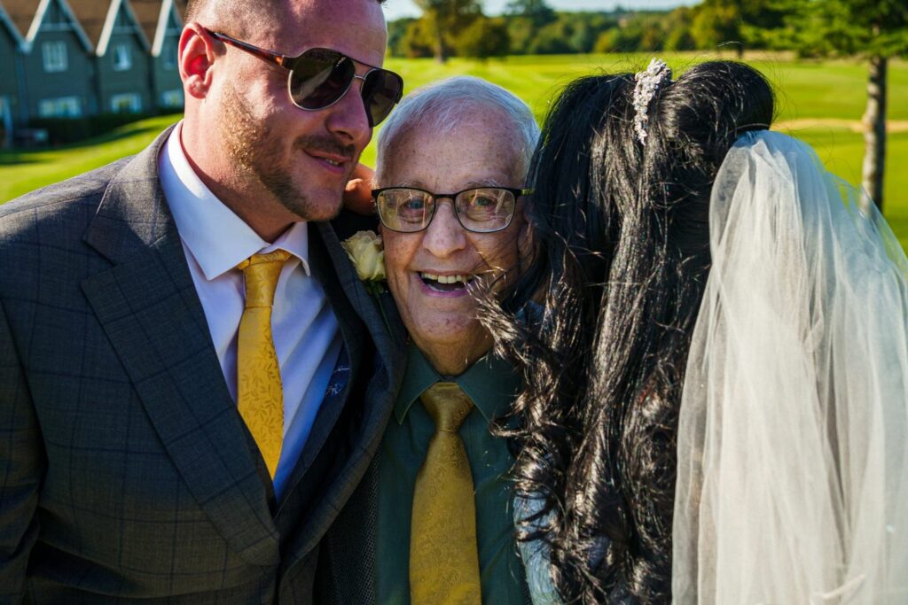 A groom in a grey suit and yellow tie, an elderly man in glasses and a yellow tie, and a bride in a white veil share a joyful moment outdoors in a sunny setting, presumably at a wedding. Image by Documentary Wedding Photographer Lewis Bishop Photography.