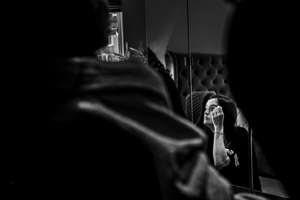 In a black and white photo, a person is seen sitting and applying makeup while looking into a mirror. The foreground features out-of-focus shoulders framing the scene, and the background shows a cushioned headboard and a modern, stylishly designed room. Image by Documentary Wedding Photographer Lewis Bishop Photography.