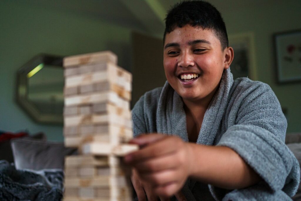 A smiling person in a gray robe is sitting on a couch, engaging in a game of Jenga. They are placing or removing a block from the middle of the tower. The setting appears to be a cozy living room with muted lighting. Image by Documentary Wedding Photographer Lewis Bishop Photography.