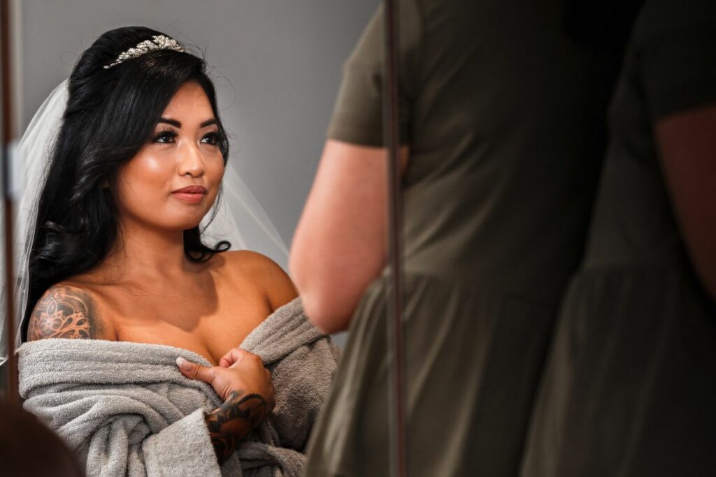A bride with a tattoo on her shoulder stands wrapped in a gray towel, looking at herself in a mirror. She wears a delicate headpiece and veil, with dark, curled hair cascading down. A person in green is reflected in the mirror, standing next to her. Image by Documentary Wedding Photographer Lewis Bishop Photography.