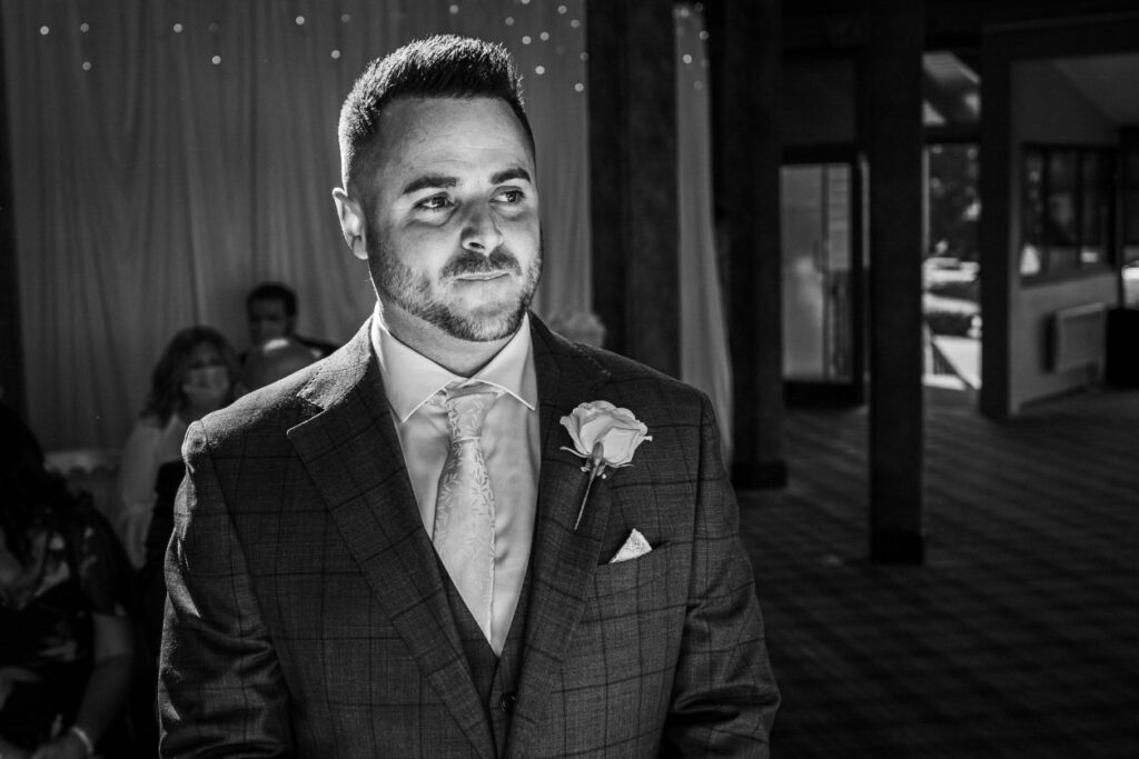 A man in a suit stands in a well-lit room, looking ahead with a thoughtful expression. He has neatly styled hair and a beard, and wears a boutonnière with a white rose on his lapel. People are seated behind him, with soft lighting creating a warm ambiance. Image by Documentary Wedding Photographer Lewis Bishop Photography.