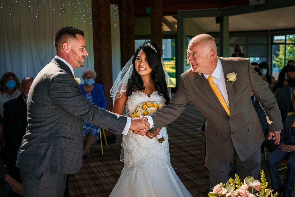 A bride in a white wedding gown and veil smiles as she stands between a man in a gray suit who is shaking hands with another man in a tan suit holding a bouquet. They are surrounded by guests, some seated and some wearing masks, in a well-lit venue. Image by Documentary Wedding Photographer Lewis Bishop Photography.