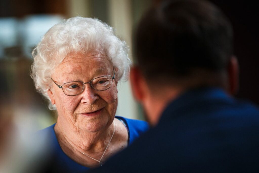 An elderly woman with white curly hair and glasses is smiling and looking past someone with short dark hair and a beard. She is wearing a blue top and a delicate necklace. The background appears to be indoors and slightly blurred. Image by Documentary Wedding Photographer Lewis Bishop Photography.