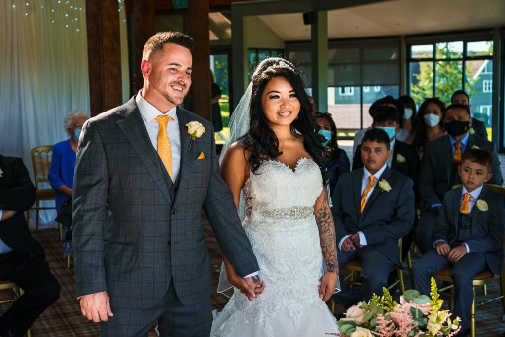 A couple stands hand in hand at their wedding ceremony. The groom is wearing a grey suit with a yellow tie, and the bride is in a white lace dress with a veil. Guests, some wearing masks, are seated and watching the couple. Flowers are arranged in the foreground. Image by Documentary Wedding Photographer Lewis Bishop Photography.