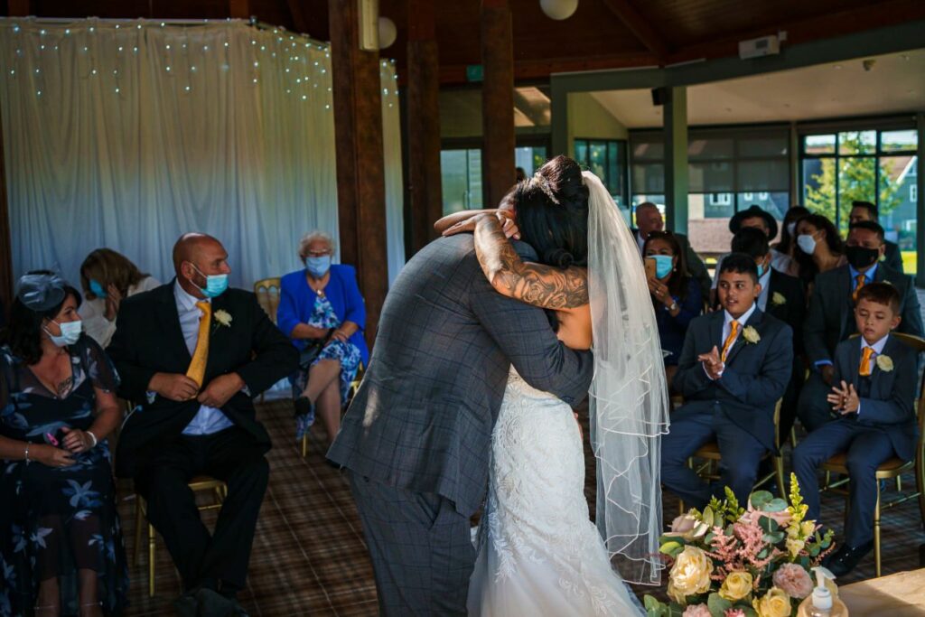 A bride and groom embrace at their wedding ceremony. The bride wears a white gown and veil, the groom a gray suit. Guests wearing masks are seated, observing the moment. Chairs are arranged in rows, and flowers are visible in the foreground. Image by Documentary Wedding Photographer Lewis Bishop Photography.