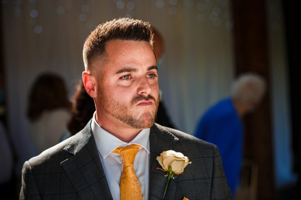 A man with short brown hair and a trimmed beard is dressed in a gray suit and a bright yellow tie with a white rose boutonniere. He is standing indoors with people blurred in the background and looking to the side under soft lighting. Image by Documentary Wedding Photographer Lewis Bishop Photography.