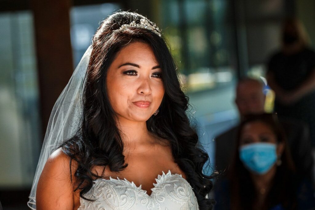 A bride with dark, wavy hair wearing a white lace wedding dress and a tiara smiles softly. She has a veil draped over her back, and there are guests in the background, some of whom are wearing face masks. The sunlight highlights her face. Image by Documentary Wedding Photographer Lewis Bishop Photography.