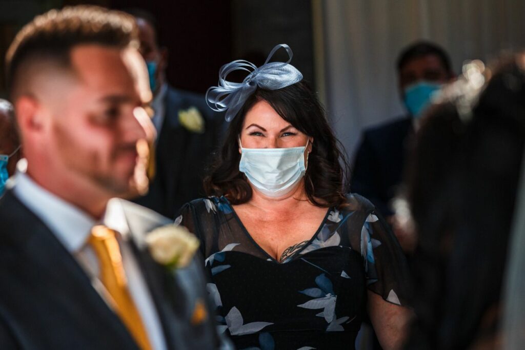 A woman dressed in a dark floral dress and a small fascinator, wearing a face mask, is smiling and looking forward at a formal event. A man in a suit with a yellow tie and boutonnière stands in the foreground, also wearing a mask. Image by Documentary Wedding Photographer Lewis Bishop Photography.