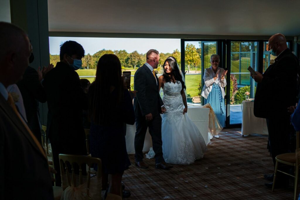 A bride in a white gown and a groom in a gray suit walk down the aisle, smiling and holding hands. Guests on either side stand and applaud, some taking photos. The background reveals an outdoor golf course visible through large windows. Image by Documentary Wedding Photographer Lewis Bishop Photography.