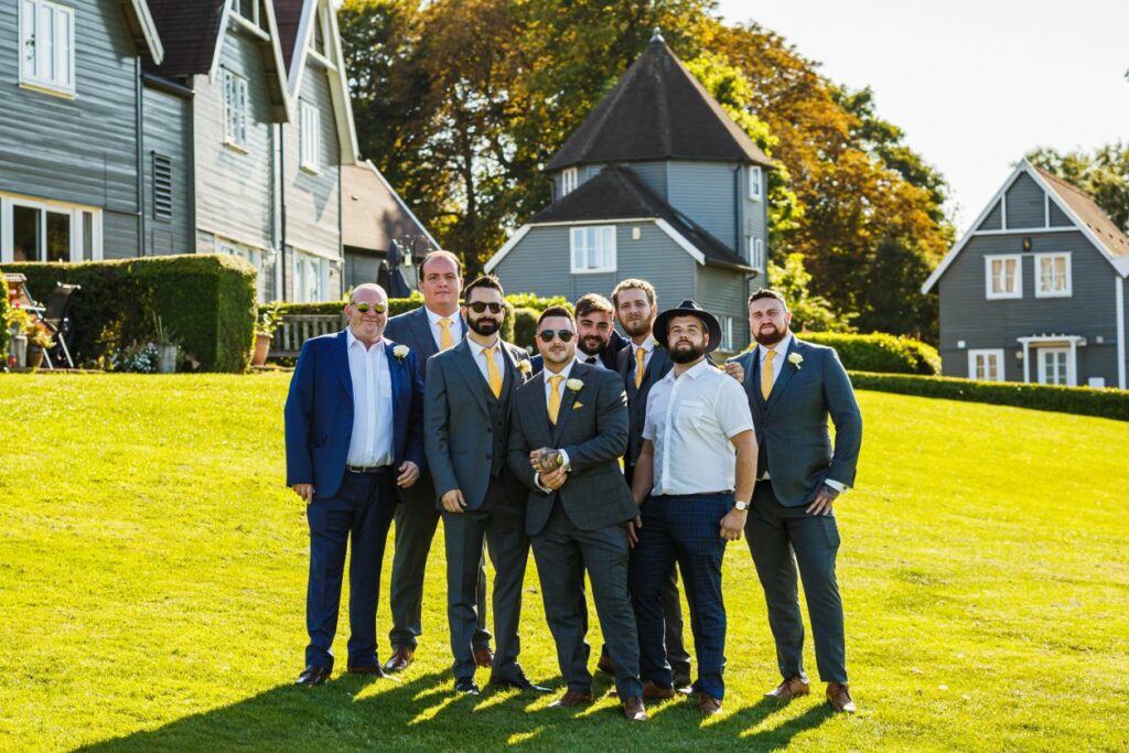 A group of seven men, dressed in suits and standing on a grassy lawn, pose for a photo on a sunny day. Some have sunglasses, and one is holding a small dog. Behind them, there are charming grey buildings with white trim and lush greenery. Image by Documentary Wedding Photographer Lewis Bishop Photography.