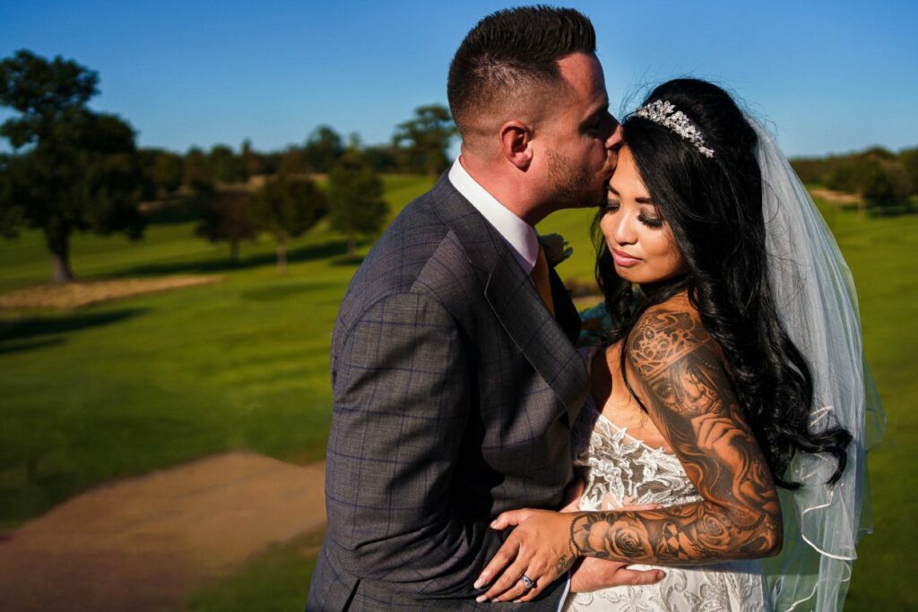 A groom in a gray suit tenderly kisses his bride, who is wearing a white lace wedding gown and a veil, on the forehead. The bride has long dark hair and visible tattoos on her arm. They stand in a green outdoor setting with trees and a clear blue sky in the background. Image by Documentary Wedding Photographer Lewis Bishop Photography.
