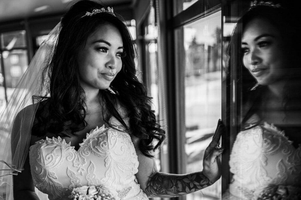 Black and white photo of a bride in a lace wedding gown with a veil, looking out a window. She is holding a bouquet and has wavy hair, with a tiara on her head. The reflection of her face is visible in the window glass. Image by Documentary Wedding Photographer Lewis Bishop Photography.