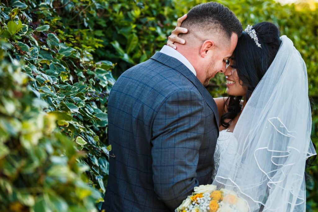 A bride and groom share an intimate moment, touching foreheads and smiling, amidst lush greenery. The bride, in a white gown and veil, holds a bouquet of white and yellow flowers, while the groom wears a gray plaid suit. The scene is bright and filled with natural light. Image by Documentary Wedding Photographer Lewis Bishop Photography.