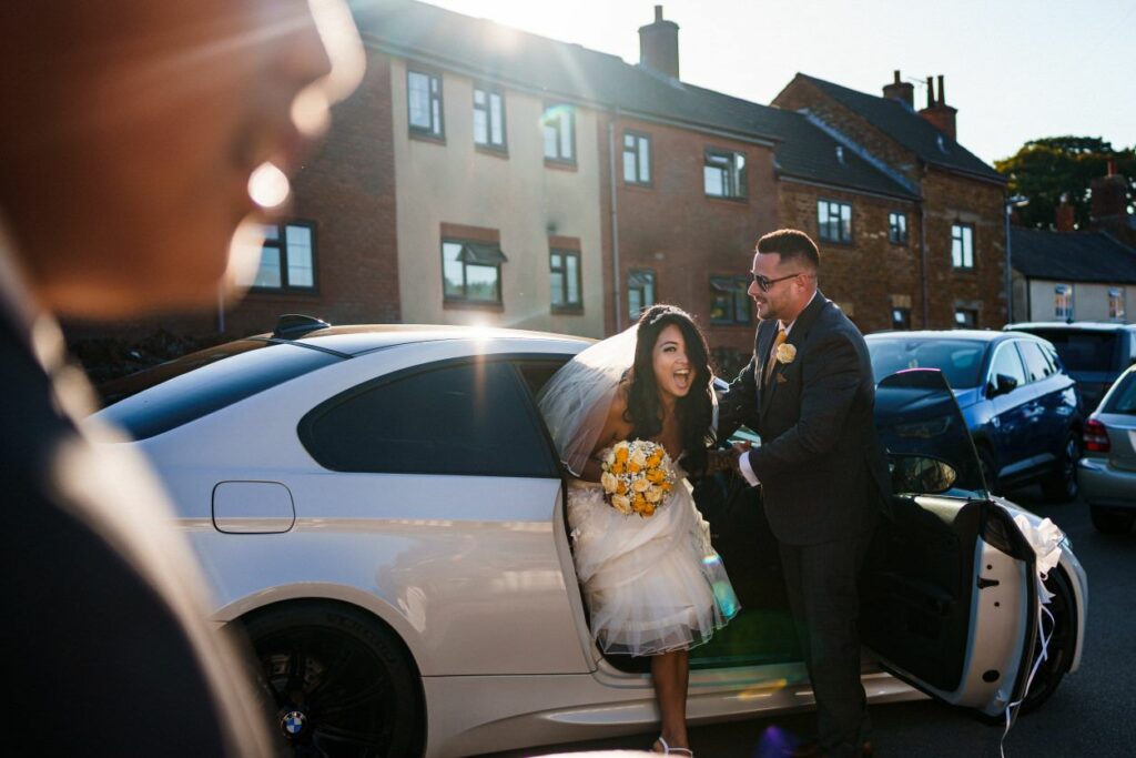A bride, wearing a veil and white dress, steps out of a white car with a joyous smile. She holds a bouquet of yellow flowers. A groom, dressed in a gray suit, offers his hand to help her. The sun shines brightly, and brick buildings are visible in the background. Image by Documentary Wedding Photographer Lewis Bishop Photography.