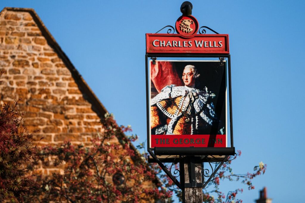 A pub sign with the name "Charles Wells" displayed at the top and "The George Inn" at the bottom. The sign features an image of a regal man in historical attire. The background includes part of a stone building and some foliage. Image by Documentary Wedding Photographer Lewis Bishop Photography.