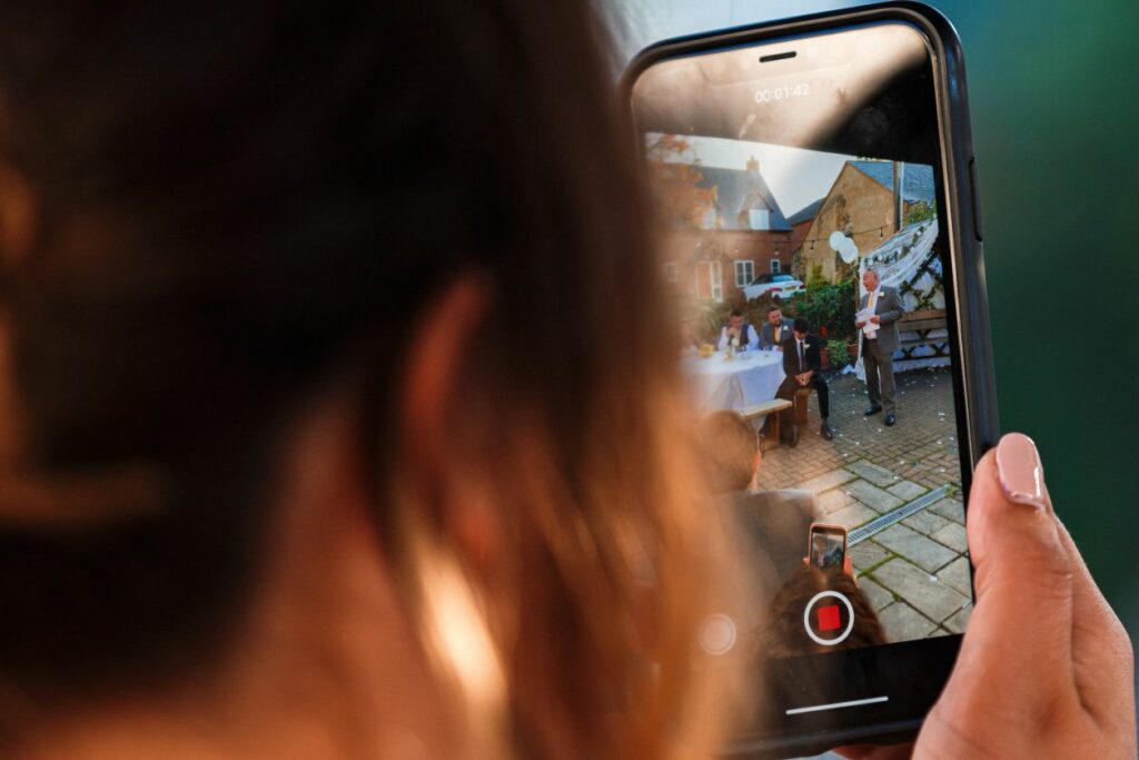 A person holds up a smartphone recording a live event, visible on the phone's screen. In the scene, people are gathered outdoors around a table with a speaker standing in the background. The setting appears to be a patio with brick flooring and a building behind. Image by Documentary Wedding Photographer Lewis Bishop Photography.