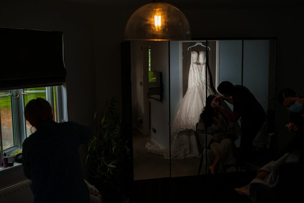 A bride gets her makeup done in a dimly lit room with a wedding dress hanging in the background. A lightbulb hangs from the ceiling, casting a warm glow. Two people are visible, one assisting with makeup and the other looking out a window. Image by Documentary Wedding Photographer Lewis Bishop Photography.