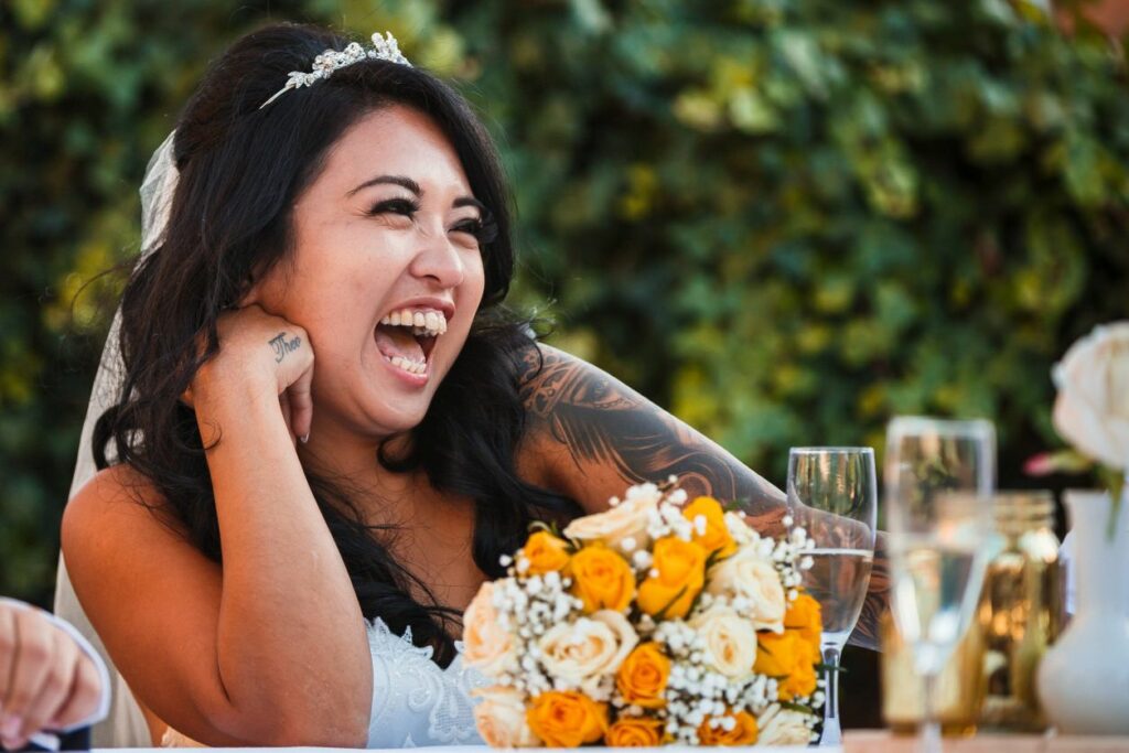 A joyful bride with long dark hair and a tiara smiles widely while resting her chin on her hand. She has visible tattoos on her arm and sits at a table adorned with a bouquet of yellow and white roses, with green foliage in the background. Image by Documentary Wedding Photographer Lewis Bishop Photography.