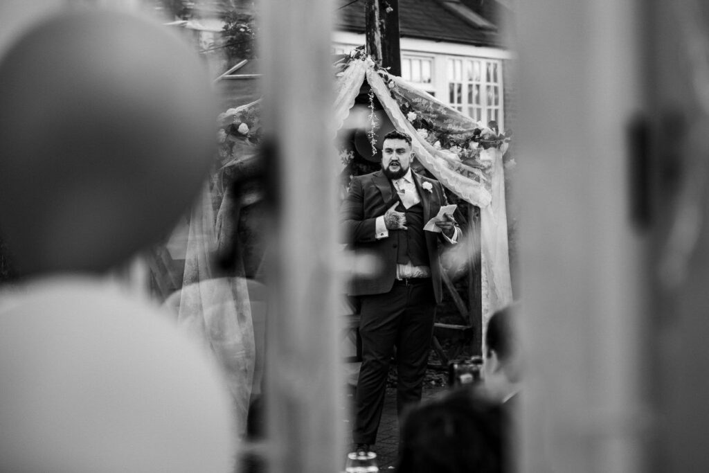 A man in a suit and bow tie gives a speech outdoors, standing under a decorated arch with tulle and flowers. The scene is viewed through a blurred foreground of what appears to be balloons, with people observing from the background. The image is in black and white. Image by Documentary Wedding Photographer Lewis Bishop Photography.