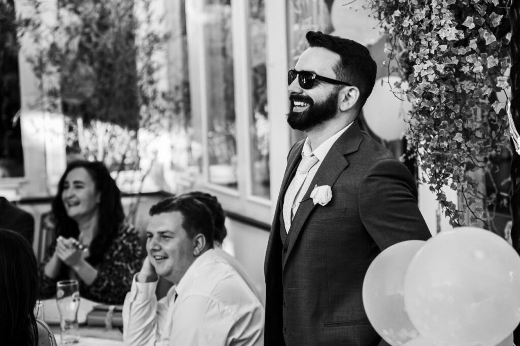 A bearded man in a suit and sunglasses is standing and laughing at an event, with a boutonnière on his lapel. He is surrounded by smiling guests seated at tables, with balloons and flowers visible in the background. The setting appears festive and cheerful. Image by Documentary Wedding Photographer Lewis Bishop Photography.
