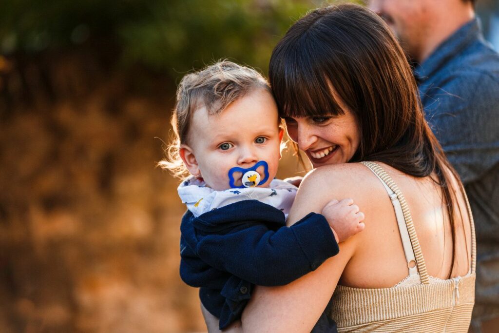 A woman with dark hair, smiling and holding a baby with a pacifier, wearing a white and navy blue outfit. The baby is looking at the camera with a curious expression. A blurred figure in the background suggests an outdoor setting. Image by Documentary Wedding Photographer Lewis Bishop Photography.