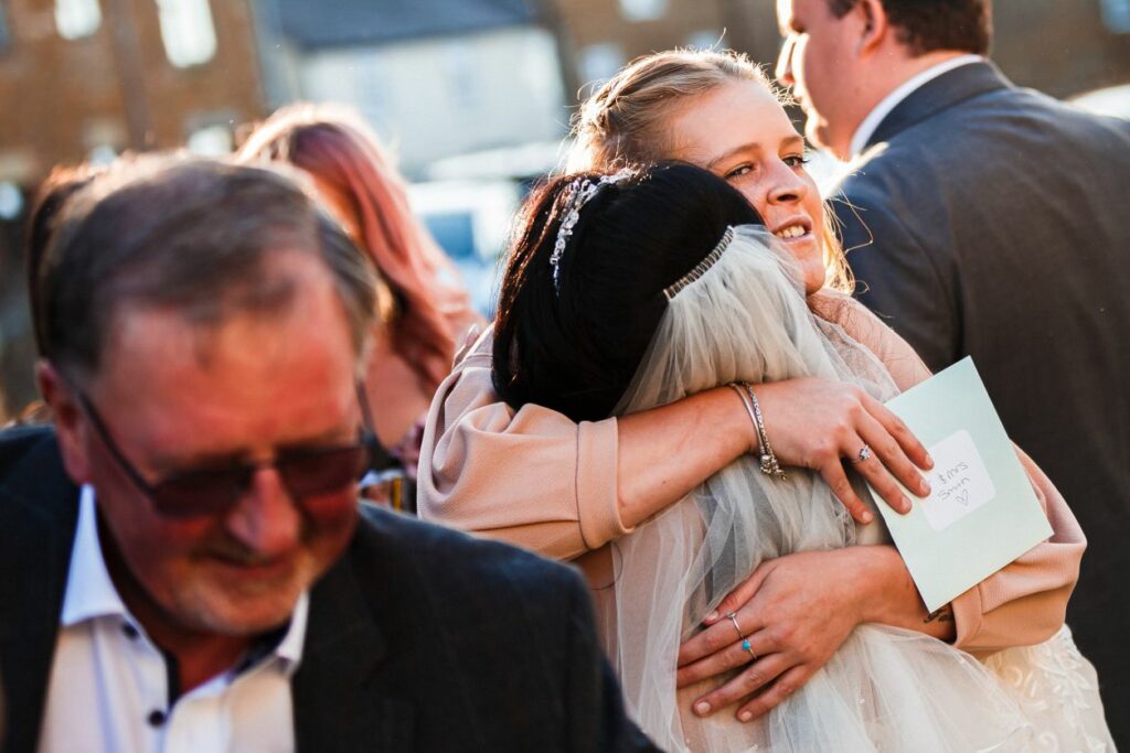 A bride in a white dress and veil hugs a woman holding an envelope, surrounded by people in formal attire. The scene appears to be outdoors on a sunny day. In the foreground, a man in sunglasses walks by. Image by Documentary Wedding Photographer Lewis Bishop Photography.