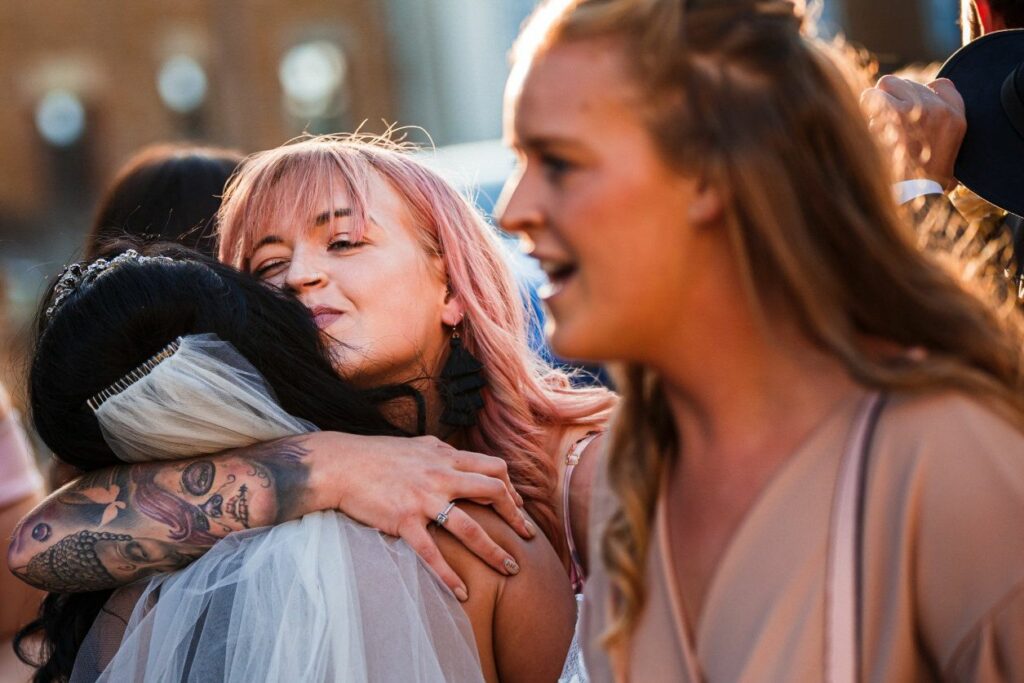 A woman with pink hair and tattoos warmly hugs a bride wearing a white veil. Another woman with long hair stands in the foreground, smiling and looking towards the hugging women. The scene appears to be at an outdoor event with blurred people in the background. Image by Documentary Wedding Photographer Lewis Bishop Photography.
