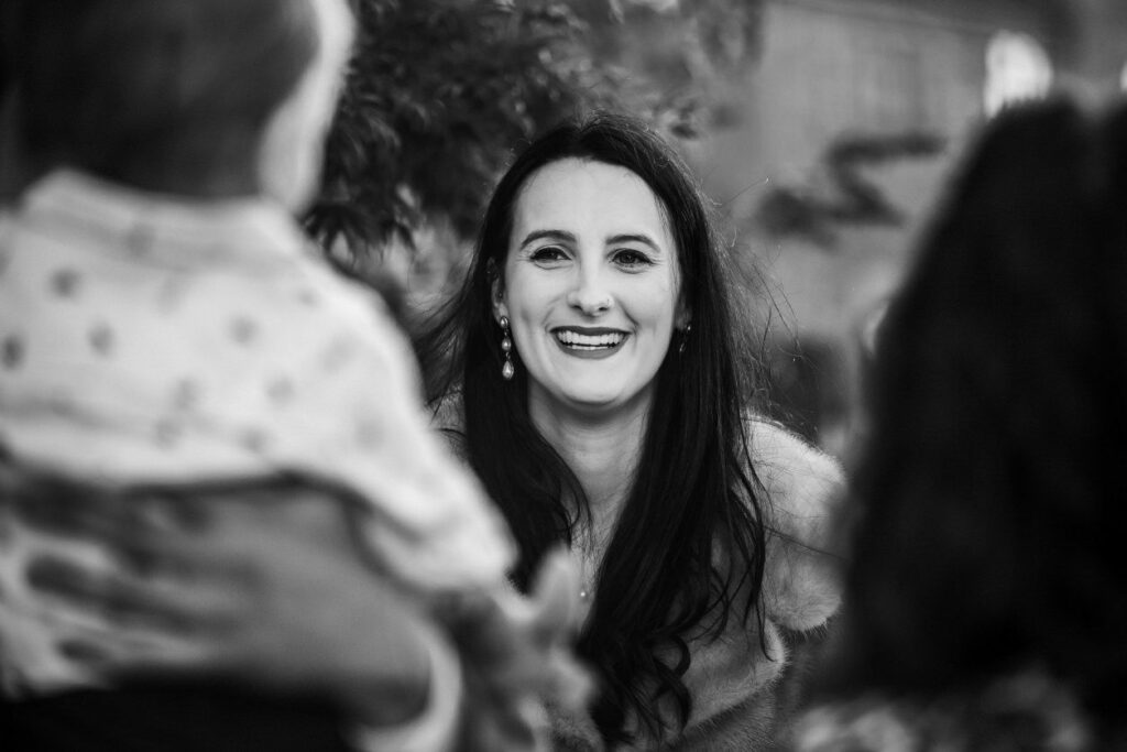 A woman with long dark hair smiles joyfully while looking at a child who is blurred in the foreground. The black-and-white photo captures a moment of happiness and connection. Trees and a building are visible in the background. Image by Documentary Wedding Photographer Lewis Bishop Photography.