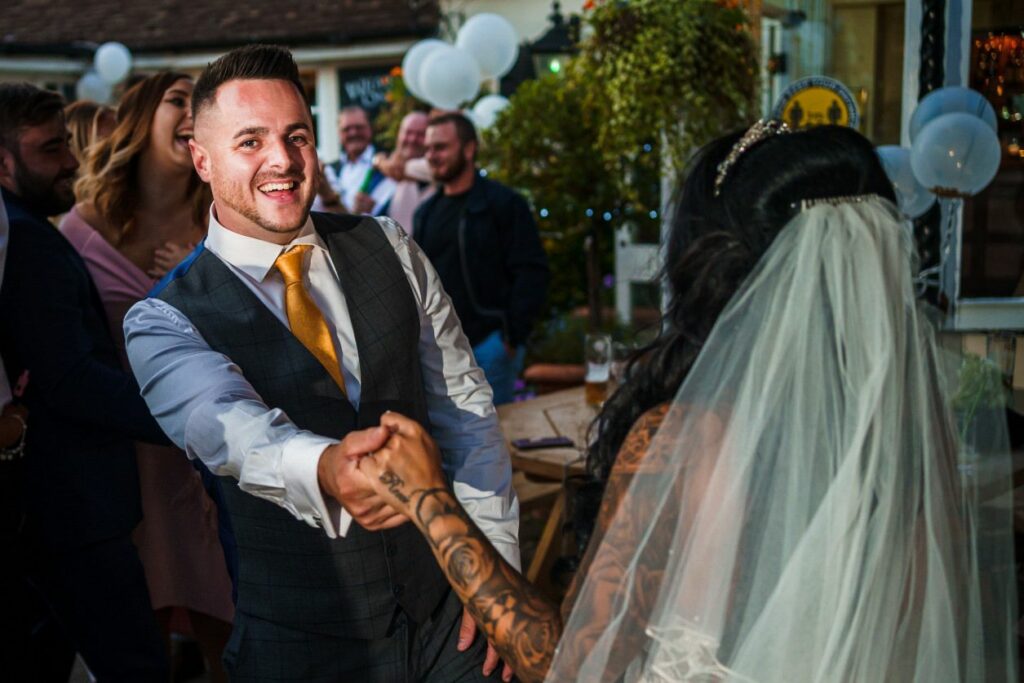 A smiling groom in a gray vest and yellow tie dances with his tattooed bride, who is wearing a white veil and a beaded headpiece. They are surrounded by guests, balloons, and colorful lights, creating a festive atmosphere. Image by Documentary Wedding Photographer Lewis Bishop Photography.