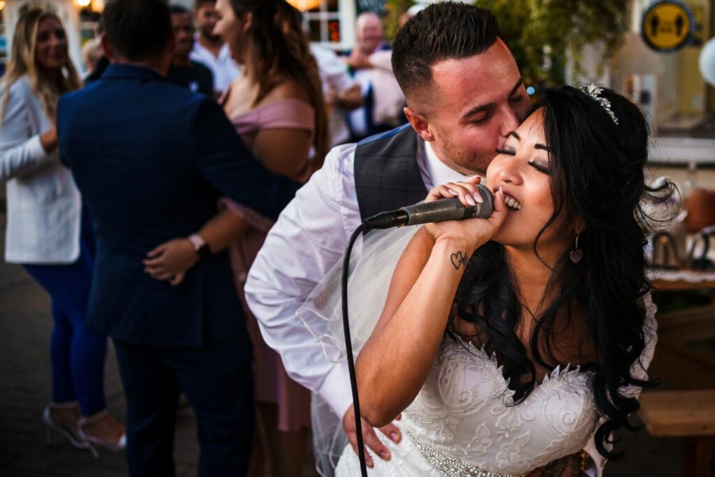 A bride in a white gown holds a microphone and sings passionately while a groom, in a gray vest and white shirt, embraces her from behind and kisses her cheek. Guests in the background are seen smiling and enjoying the moment at the wedding reception. Image by Documentary Wedding Photographer Lewis Bishop Photography.